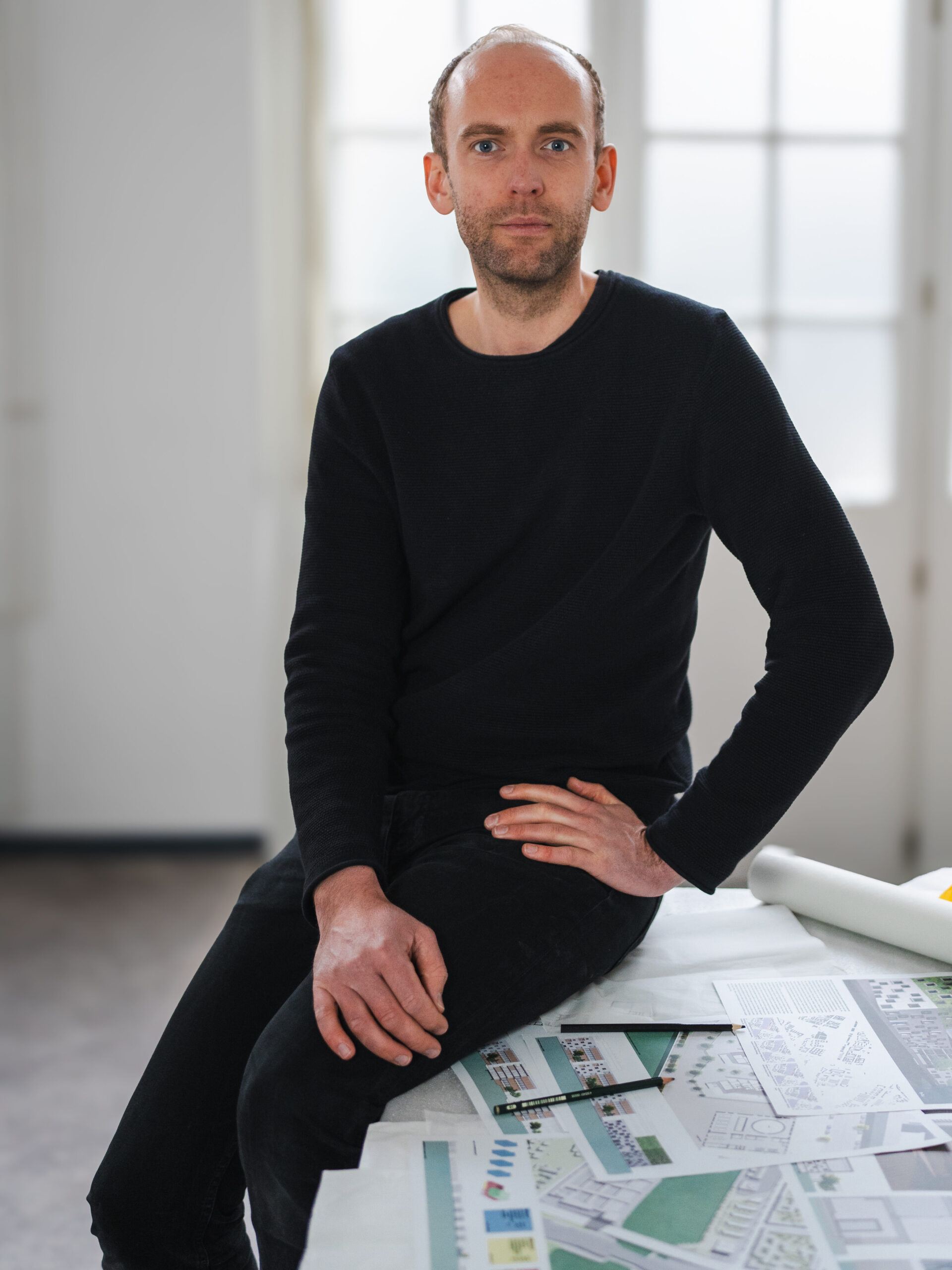 Inhaber Andreas Lang sitzt während der Arbeit im Studio auf einem Tisch, auf dem Skizzen, Baupläne und ein Bleistift liegen und schaut in die Kamera.
