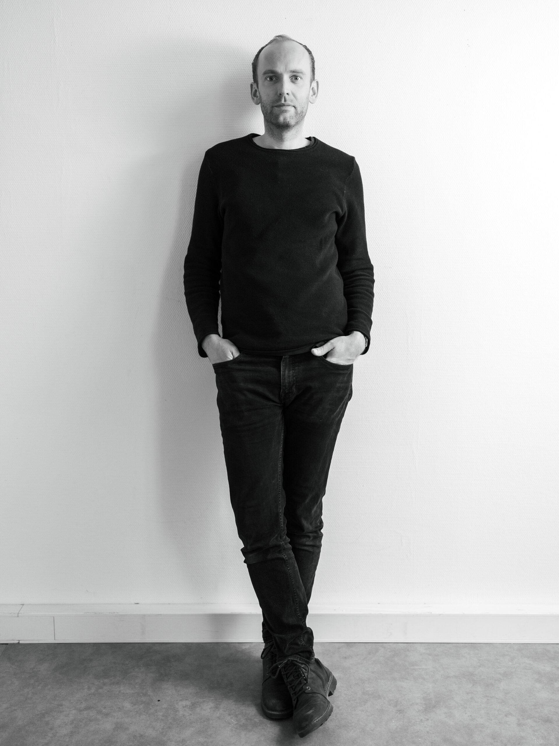 Architekt Andreas Lang steht stilvoll schwarz gekleidet mit den Händen in den Hosentaschen vor einer weißen Wand und schaut freundlich interessiert in die Kamera.