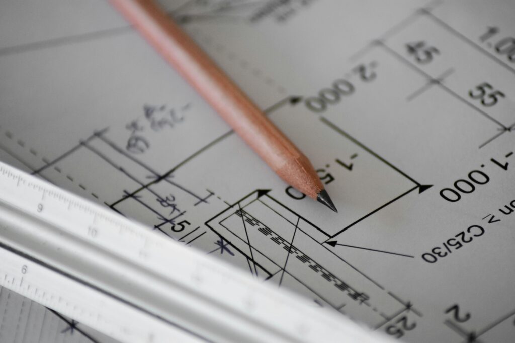 Ein Bleistift und ein Dreikantmaßstab liegen auf einem Bauplan, in dem handschriftliche Notizen zu erkennen sind.