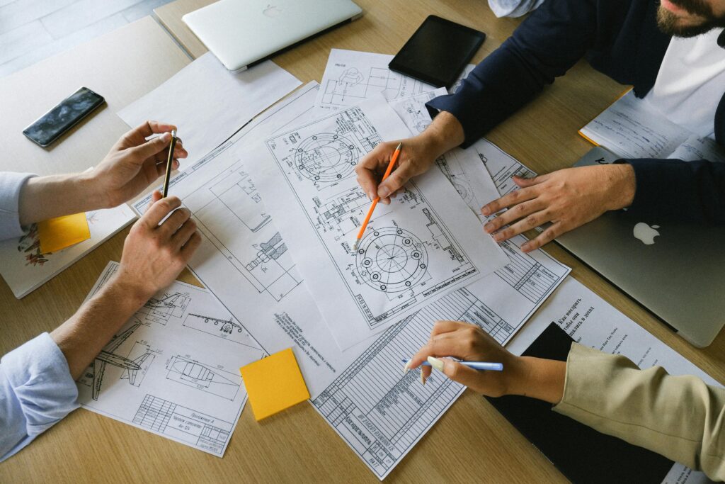 Zwei Menschen sitzen zusammen mit dem Architekten an einem Tisch, auf dem Baupläne verteilt sind und beraten über die ideale Projektstruktur für ihr anstehendes Bauvorhaben.