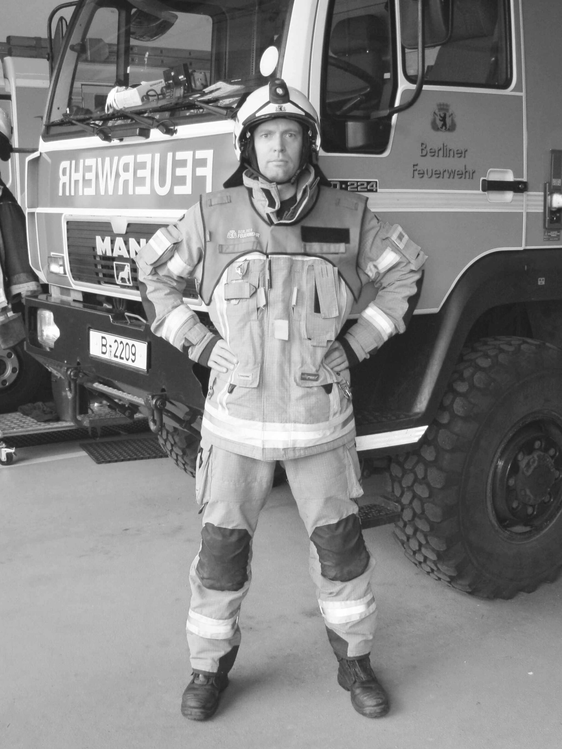 Andreas Lang steht in Feuerwehrschutzausrüstung mit Helm und Stiefeln vor einem Feuerwehrfahrzeug und schaut entschlossen in die Kamera.