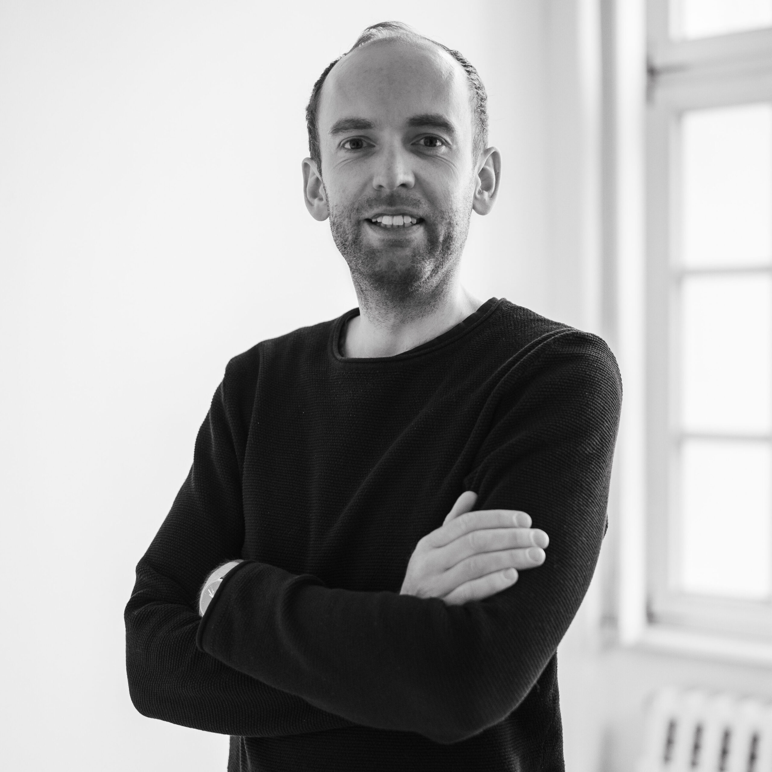Inhaber Andreas Lang steht im Studio mit verschränkten Armen vor einer weißen Wand mit Fenstern und schaut freundlich und entschlossen in die Kamera.