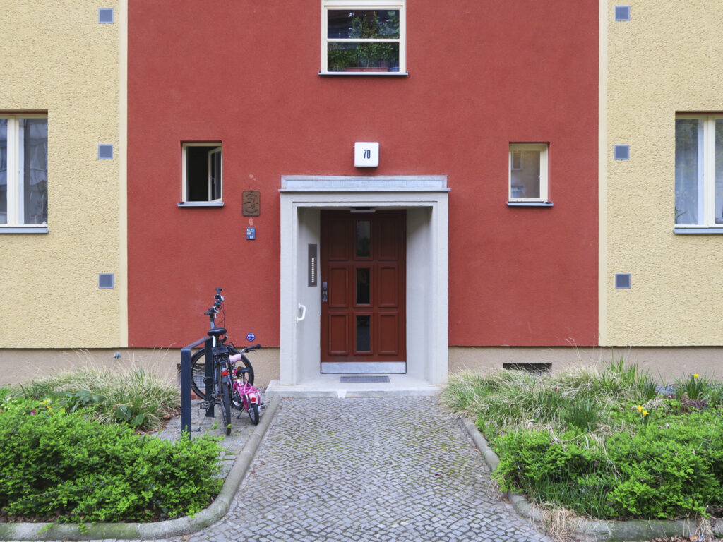 Blick auf einen Hauseingang mit einer braunen hölzernen Eingangstür, einem grauen steinernen Vordach sowie der charakteristischen Farbgebung der Fassade in rot und ocker.