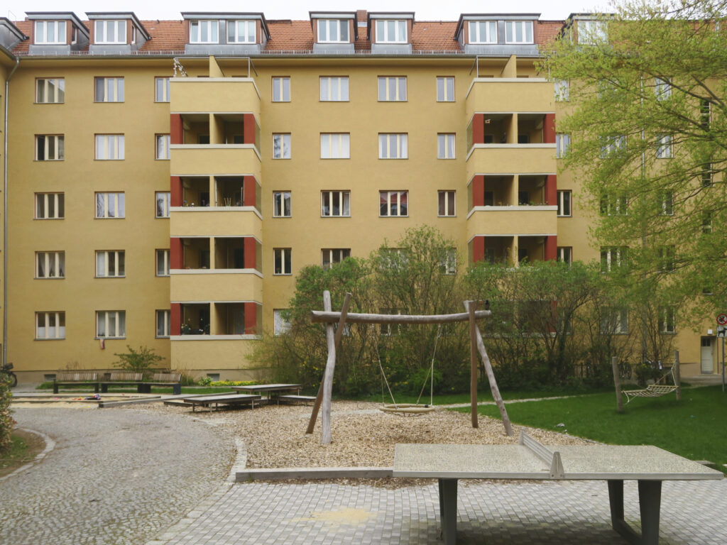 Blick in den Innenhof mit den ockerfarbenen Putzfassaden und einen Spielplatz mit Schaukel und Tischtennisplatte im Vordergrund.