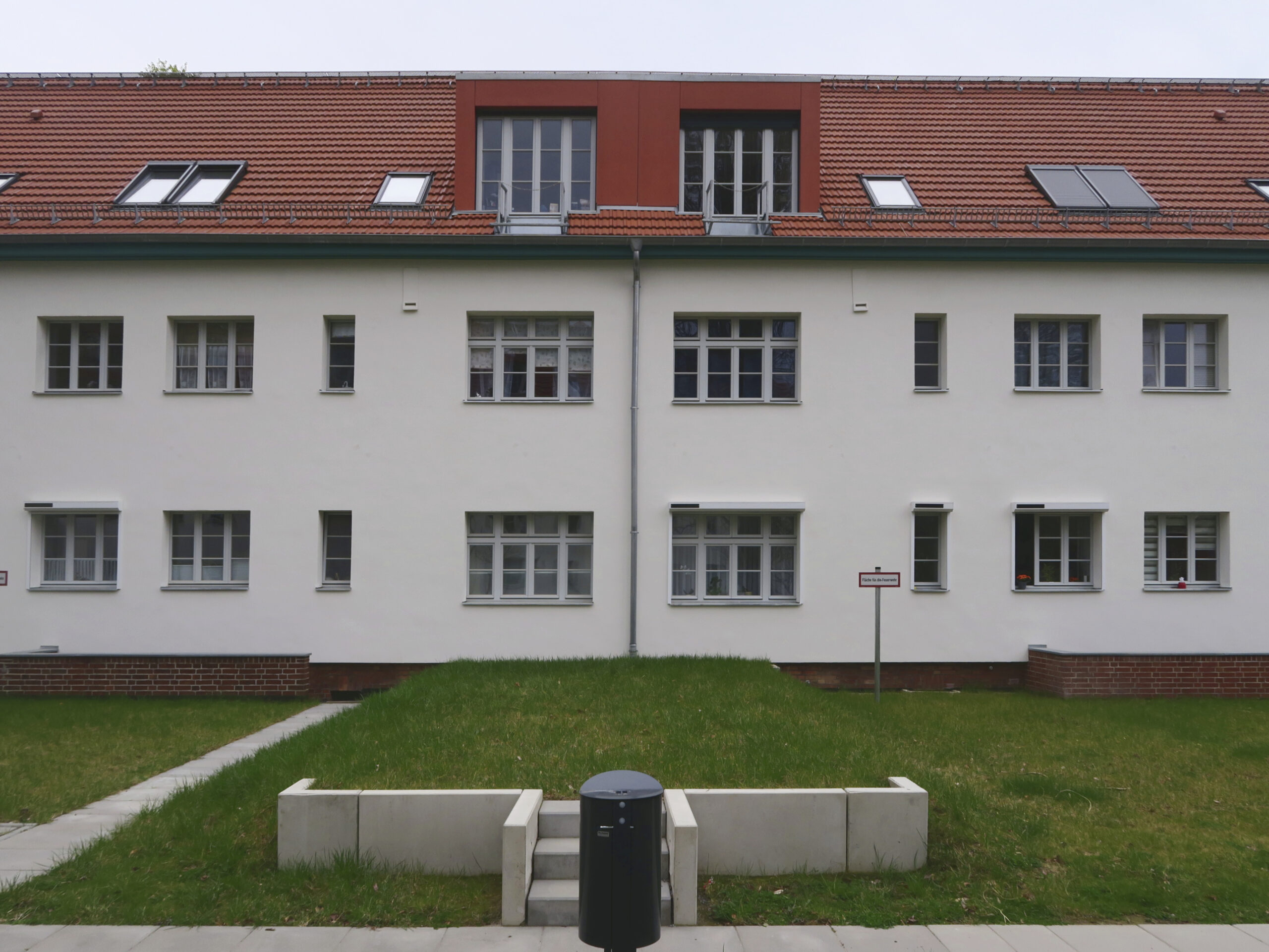 Blick auf die neu gestaltete Fassade eines zweigeschossigen Wohnhauses mit neu gebauten Dachgauben und einer Erhöhung der Geländeoberfläche zur Sicherstellung des zweiten Rettungsweges über Leitern der Feuerwehr.