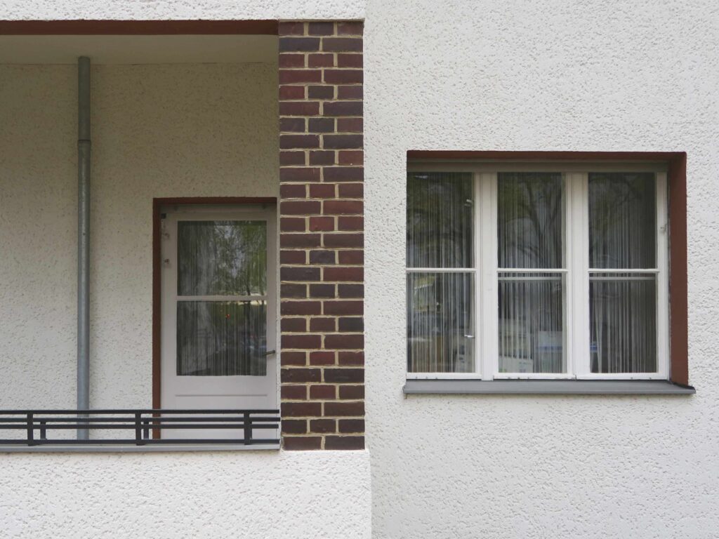 Detailaufnahme der Putzfassade und einem Fenster mit rötlich abgesetzter Laibung und einem Balkon mit restaurierten dunkelbraunen Klinkerflächen.