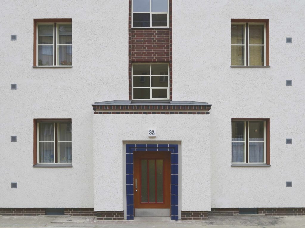 Blick auf die braune Haustür eines Hauseingangs mit roten Details und einem leuchtend blauen Rahmen aus glasierten Formsteinen.